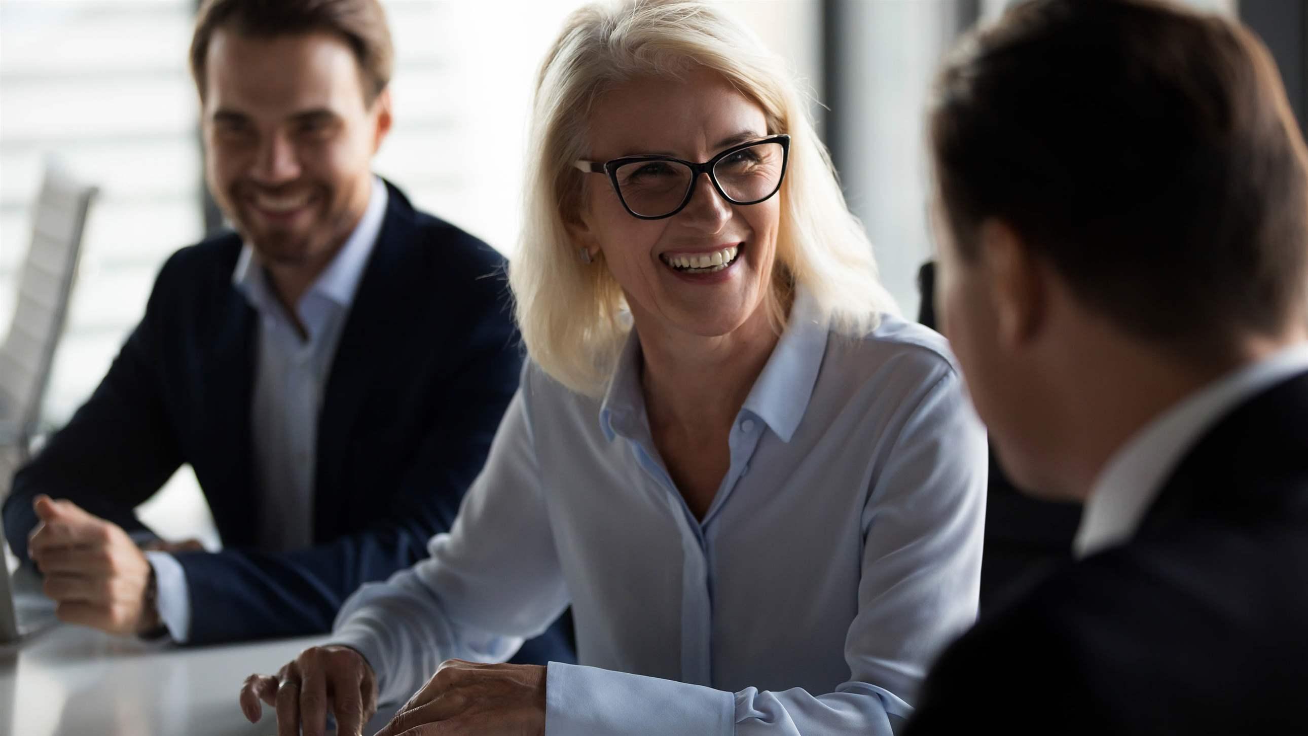 Friendly middle aged female leader laughing at group business meeting, happy old businesswoman enjoying fun conversation with partner, smiling mature business coach executive talking to colleague